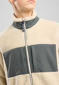 Giacca in pile di colore crema e verde scuro, con colletto alto, chiusura con zip e superficie testurizzata con pannelli lisci.
