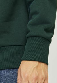 Felpa verde scuro con polsini e orlo a coste. Primo piano di una mano appoggiata su un tessuto testurizzato, che mostra i dettagli del materiale e della vestibilità.
