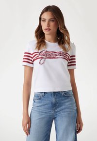 Witte cropped t-shirt met rode horizontale strepen en een geknoopt detail over de borst. Gecombineerd met lichtblauwe denim jeans.