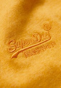 Gul stof med en blød tekstur, der har et broderet logo med teksten "SuperDry" samt dekorative elementer og en accent.