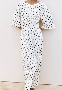 Robe blanche à pois noirs, corsage froncé et manches évasées aux coudes portée par une personne debout contre un mur clair.