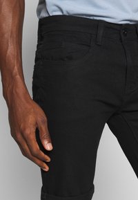 Jeans de mezclilla negra con corte slim, que cuentan con cinco bolsillos, cierre de botón y dobladillo enrollado, destacando una textura suave.