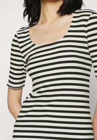 Vihreä ja valkoinen raidoitettu toppi, jossa on pyöreä pääntie ja lyhyet hihat. Väretty pinta, jossa on rispaantuneet reunat hihansuiden kohdalla.