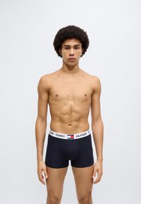 Μαύρα ανδρικά εσώρουχα boxer με λευκή μέση που φέρει το λογότυπο "TOMMY HILFIGER". Το μοντέλο έχει κοντά, σγουρά μαλλιά και μυώδες σώμα.