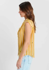 Femme aux longs cheveux bruns portant un chemisier sans manches jaune et blanc à rayures avec de petits détails floraux et un jean bleu clair, debout de profil.