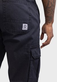 Dunkle Cargo-Hosen aus Baumwolle, mit seitlichen Taschen und einem Markenschild an der Gesäßtasche. Der Stoff hat eine glatte Textur.