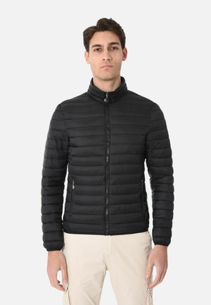 Uomo di fronte che indossa una giacca trapuntata nera con zip e pantaloni beige chiaro su uno sfondo bianco uniforme.