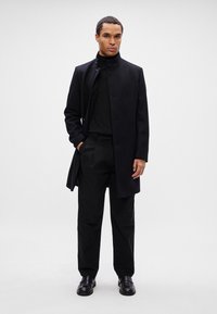 Jonge man staat, gekleed in een zwart hoogsluitend jasje, zwarte turtleneck, zwarte broeken en zwarte nette schoenen tegen een effen lichte achtergrond.
