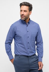 Camicia a scacchi blu e bianca con bottoni, maniche lunghe, dotata di un colletto classico e bottoni scuri, abbinata a pantaloni grigi su misura.