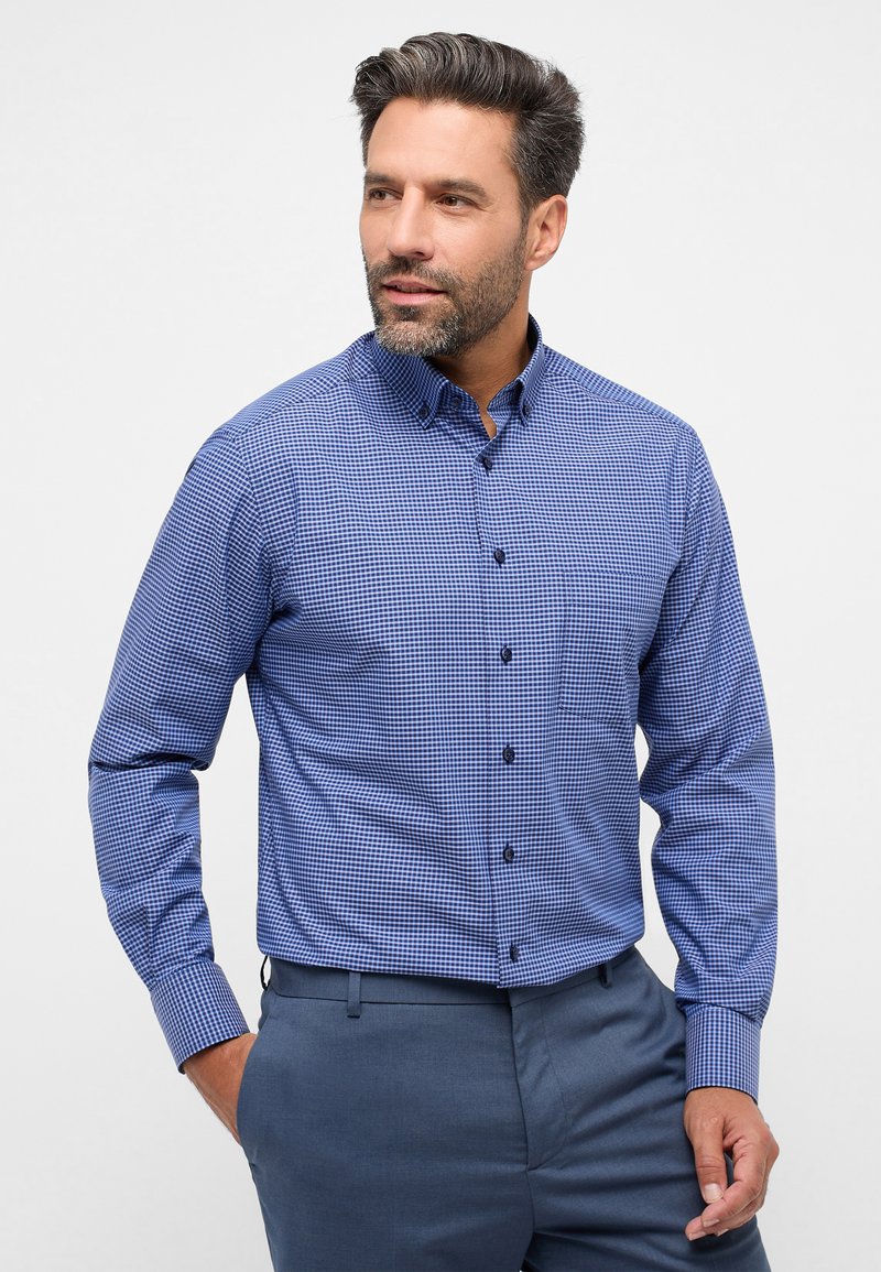 Camicia a scacchi blu e bianca con bottoni, maniche lunghe, dotata di un colletto classico e bottoni scuri, abbinata a pantaloni grigi su misura.