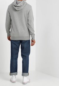 Sudadera gris con capucha ajustable, combinada con jeans oscuros. Puños enrollados visibles y zapatillas blancas de caña alta. Tejido suave y corte relajado.