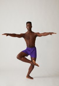 Paarse shorts met een elastische tailleband op een staande man die op één voet balanceert, met uitgestrekte armen, tegen een lichte achtergrond.
