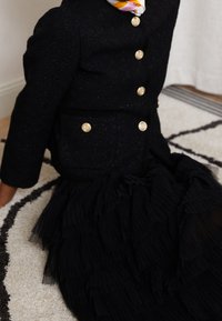 Černá strukturovaná bunda se zlatými knoflíky v kombinaci s volánkovými černými šaty. Tkanina obsahuje vrstvy a různé délky skladů.