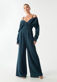 Tirkīza satīna kombinezons ar plašiem galiem, dziļu dekoltē un pūkainām piedurknēm. Iekļauta pieguļoša vidukļa daļa un plāni plecu siksnas. Gluda tekstūra.