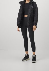 Black quilted jacket with hood, paired with a black crop top and leggings. Black sneakers with a circular logo and light-colored sole.