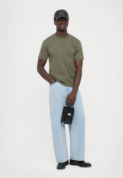 Oliven grøn kortærmet t-shirt, lyseblå løse jeans, sorte støvler og en sort kasket. Holder en lille sort taske med sølvdetaljer.