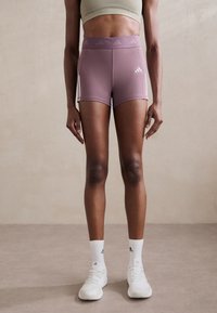 Lila sportshorts med vita sidsränder och logotyp, kombinerade med vita strumpor och skor. Materialet verkar stretchigt och mjukt.