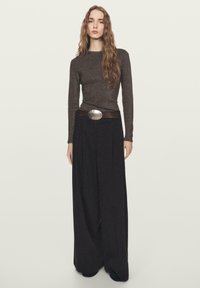 Bruine top met lange mouwen en een nauwsluitende pasvorm met een subtiele glans, gecombineerd met brede zwarte broek en een bruine riem met een ronde zilveren gesp.