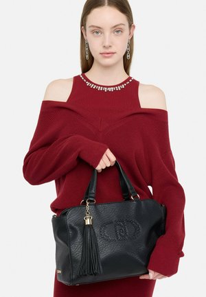 Jeune femme avec de longues boucles d'oreilles et un pull rouge à épaules dénudées tenant un sac à main en cuir noir avec un pompon et un logo clouté.