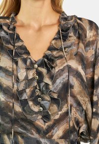 Blusa transparente con un profundo escote en V y detalles con volantes, en tonos tierra de marrón y gris, acentuada con botones metálicos en el frente.