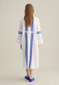 Abito lungo bianco con ricamo geometrico blu lungo il centro e le maniche. Il modello indossa scarpe basse blu. Tessuto morbido con una vestibilità comoda.
