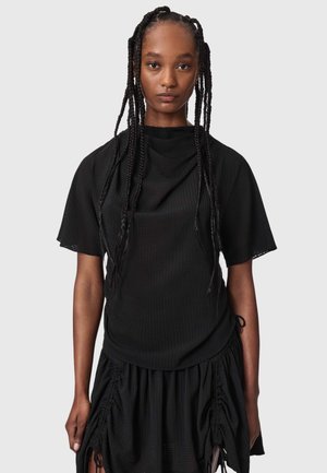Femme avec de longues tresses portant un haut noir à manches courtes et une jupe noire froncée sur un fond gris uni.