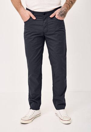 Mann trägt schwarze gerade geschnittene Hose, weiße Sneakers und ein weißes Hemd, Hände in den Taschen, linker Unterarm mit Tattoos sichtbar.