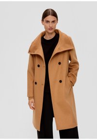 Manteau camel croisé avec un large col, fabriqué en laine texturée, avec des boutons noirs et des poches latérales, porté sur un haut noir.