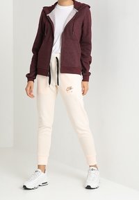 Bordeaux zip-up hoodie med fickor, krämfärgade joggers med Nike-logga och vita sneakers. Mjuk tyg, ribbade manschetter och justerbar dragsko.