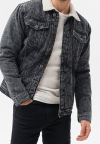 Giacca di jeans nera sbiadita con colletto in pile. Presenta tasche sul petto e chiusure con bottoni. Indossata sopra una camicia bianca a trama.