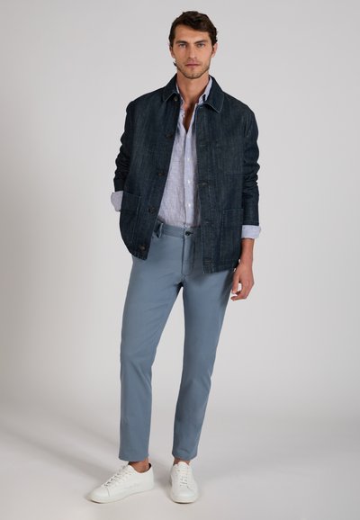 Man wearing dark denim jacket over light blue button-up shirt, blue trousers, and white sneakers standing against plain background.