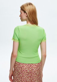 T-shirt verde chiaro a coste con bordo in pizzo sul colletto e sulle maniche, forma aderente, indossato con una gonna a fantasia caratterizzata da macchie rosse su fondo crema.