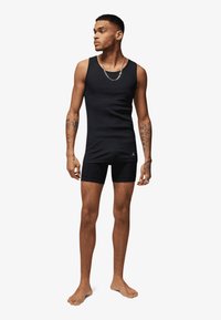 Zwart atletisch tanktop en korte broek gemaakt van rekbare stof. Voorzien van een ronde halslijn en een klein logo op de linkerborst. Blootvoets.