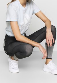 Camiseta de algodón azul claro con un logo blanco, combinada con mallas texturizadas en negro y gris. Zapatillas deportivas blancas completan el atuendo.