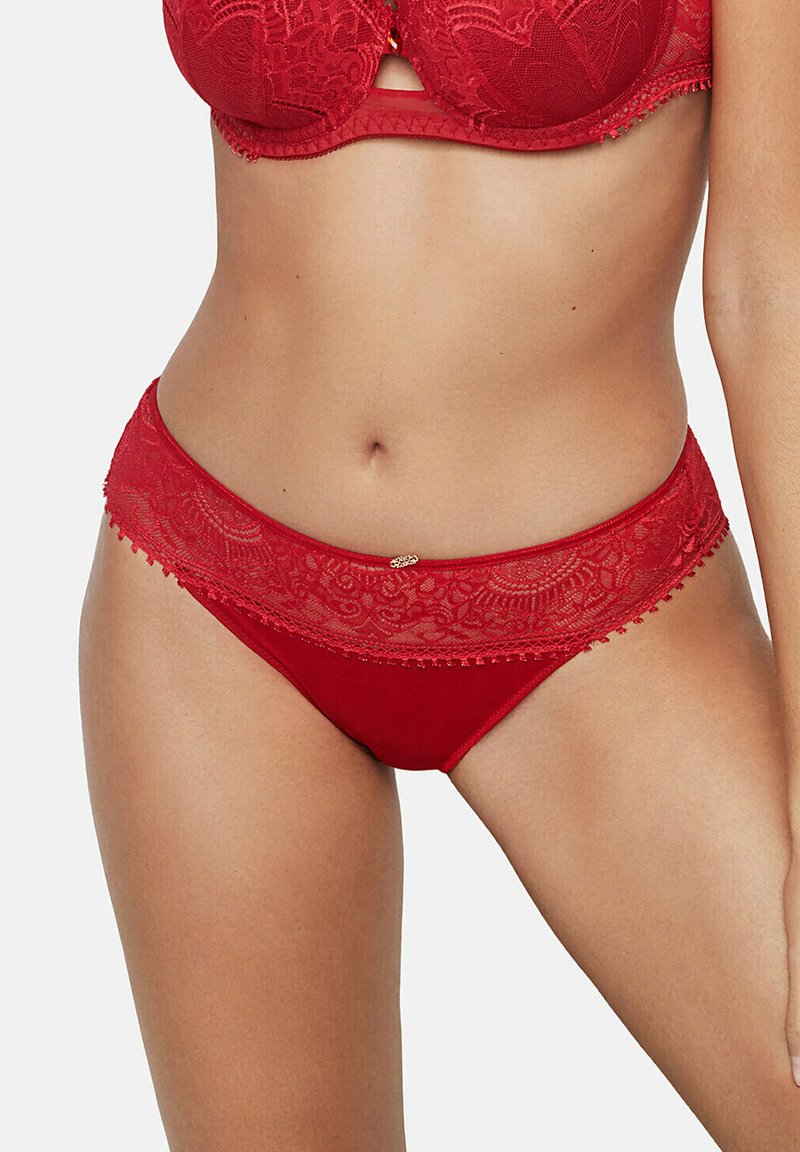 Vrouw die een rode kanten beha en bijpassende rode slip met kanten rand draagt, zichtbaar van midden torso tot bovenbenen tegen een witte achtergrond.