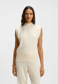 Knit sleeveless top in off-white with a high collar, ribbed hem, and smooth texture. Features minimalist design and subtle seam detailing.