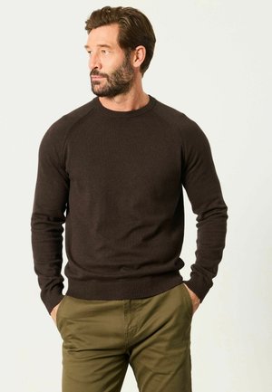 Mann mit Bart, der einen dunkelbraunen Pullover und olivgrüne Hose trägt, steht mit den Händen in den Taschen und blickt nach links vor hellem Hintergrund.