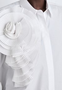 Camicia bianca con un ampio dettaglio in riga texturizzata sulla spalla, realizzata in tessuto plissettato, con un colletto sottile e chiusura a bottoni.