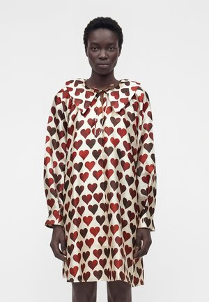 Kobieta w kremowej sukience z długim rękawem, pokrytej wzorami czerwonych i brązowych serc, z dużym spiczastym kołnierzem zawiązanym na szyi.