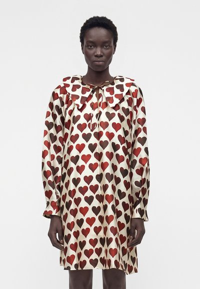 Mujer con un vestido crema de manga larga cubierto de patrones de corazones rojos y marrones con un gran cuello puntiagudo atado al cuello.