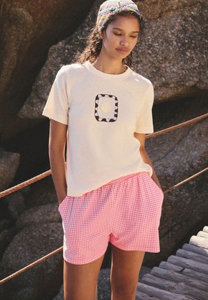 Fiatal nő fehér pólóban, amelyen kagylóminta látható, és rózsaszín kockás rövidnadrágban, egy fapályán áll kövek közelében.