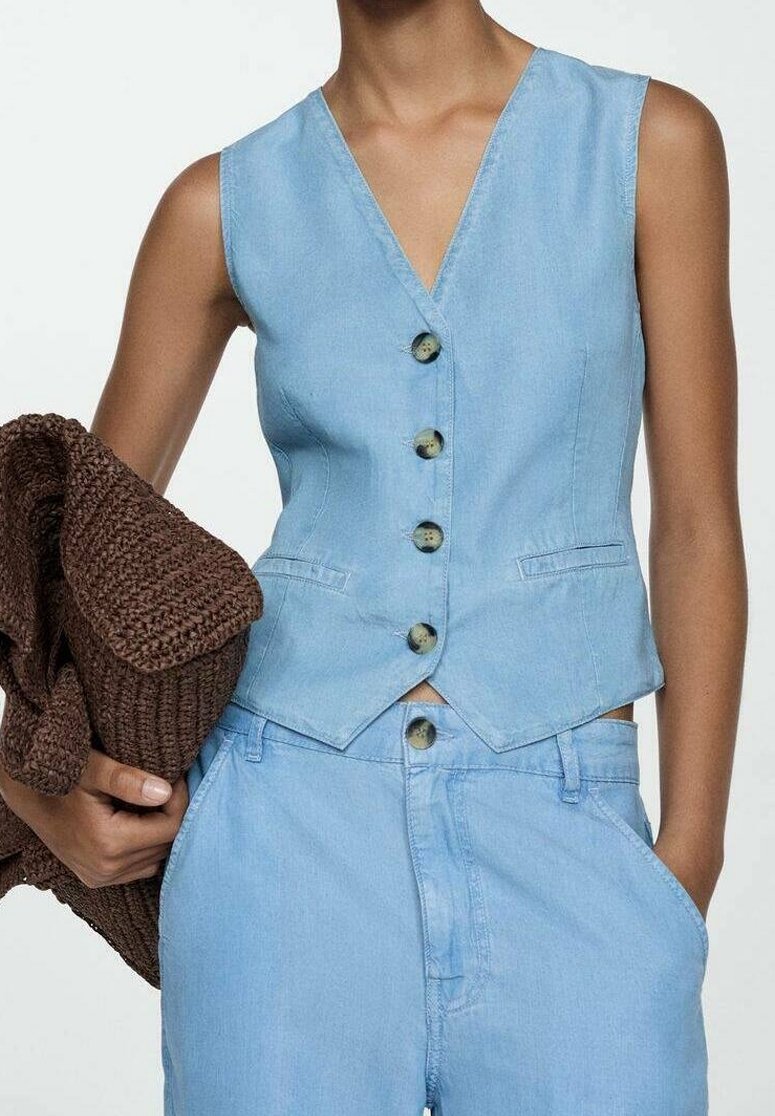 Veste en denim bleu clair avec un col en V, six boutons sur le devant et deux poches latérales. Associée à un pantalon bleu clair assorti et un châle en tricot marron.