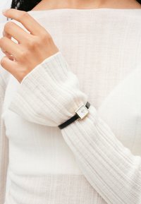 Black leather bracelet with a gold square watch face featuring a white dial, worn on a hand with a light, textured sweater.
