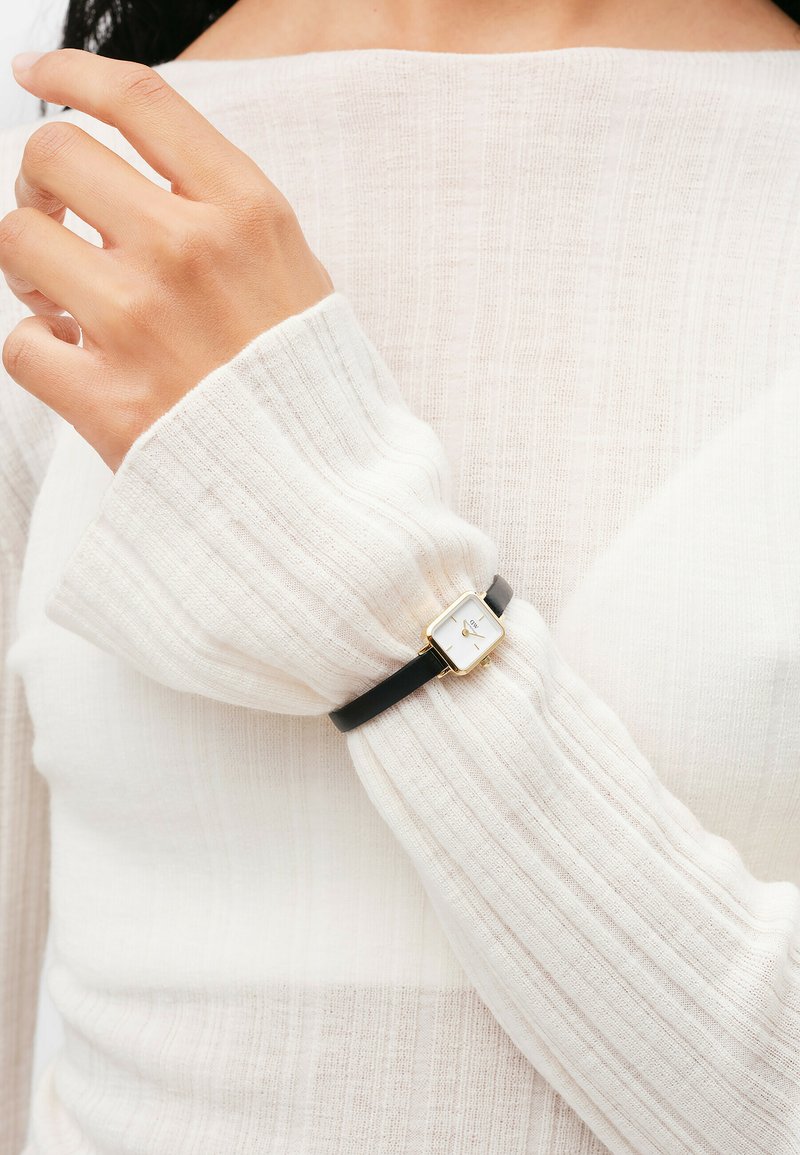 Black leather bracelet with a gold square watch face featuring a white dial, worn on a hand with a light, textured sweater.