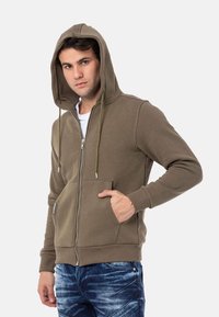 Olijfgroen ritsjack met een trekkoordkap, zijzakken, ribgebreide manchetten en een gladde textuur, gedragen over een wit shirt.