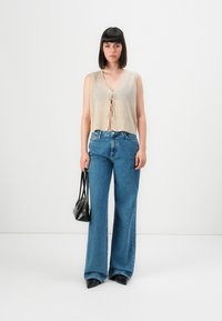Vrouw in een mouwloze beige gebreide top met voorbinding, brede blauwe jeans, zwarte puntige schoenen, met een kleine zwarte handtas, staand op een witte achtergrond.