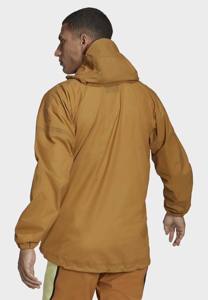 Hombre con una chaqueta con capucha de color mostaza y pantalones a juego, visto desde atrás contra un fondo claro.