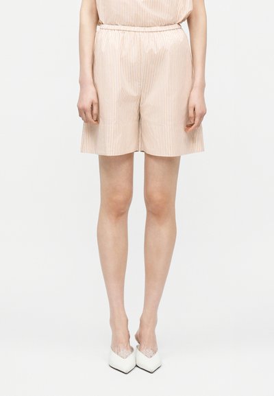 Femme portant un short beige et blanc à rayures verticales avec une ceinture élastique et des chaussures blanches à bout pointu, debout devant un fond blanc.