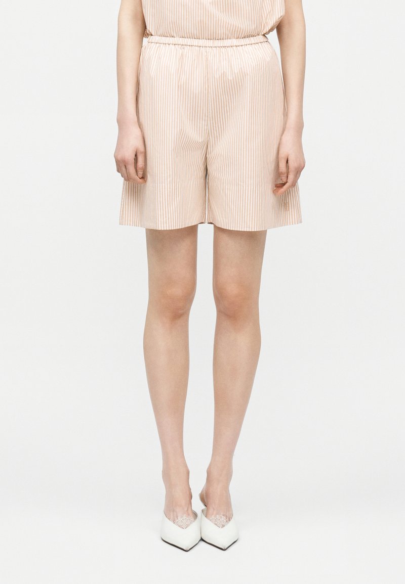 Žena nosiaca béžové a biele kraťasy s vertikálnymi pruhmi a elastickým pásom, obutá v bielych topánkach so špicatou špičkou, stojaca pred bielym pozadím.