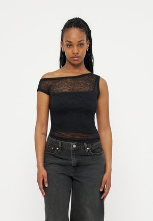 Femeie cu păr lung împletit, purtând un top negru asimetric din dantelă, cu umerii goi, și o pereche de blugi gri închis, cu talie înaltă, stând în picioare pe fundal alb.